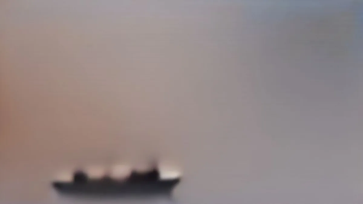 A naval vessel cruising through coastal waters at twilight near industrial energy infrastructure.