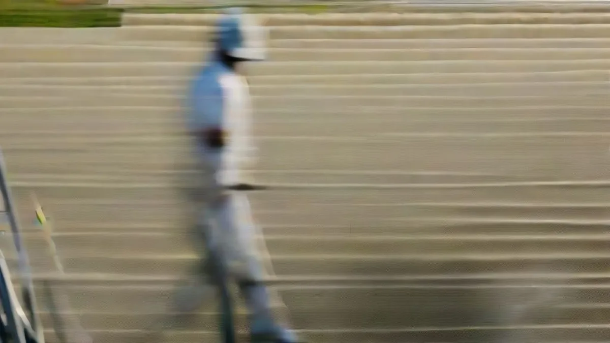 A cricket batsman walking near the boundary on a sunlit cricket field with batting pads and gloves